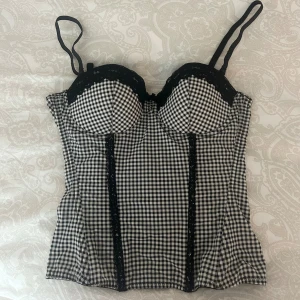 Rutig korsett med spets - Supersnygg korsett med svartvitt rutigt mönster och svarta spetsdetaljer längs bysten och framtill. Justerbara axelband och hakknäppning bak. Är väldig stretchig, jag har A/B kupa passar mig men hade absolut passat någon med större byst med ❣️❣️