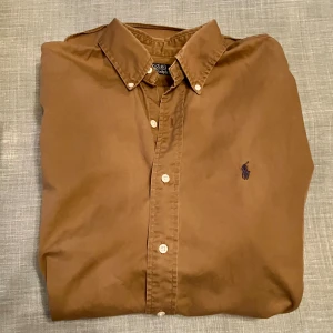 Brun skjorta från Polo Ralph Lauren - Snygg brun skjorta från Polo Ralph Lauren med klassisk krage och knappar framtill. Skjortan har lång ärm och det ikoniska Polo-logot broderat i blått på bröstet. Skjortan är oversized, snarare Large än Medium. Se mått på bild. Obs, bilderna med mått visar tvillingskjorta i annan färg. 