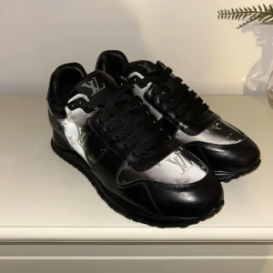  svarta och silver sneakers - Snygga sneakers i svart och silver med ikoniska LV-monogramdetaljer. Skorna har snörning, platt sula och är tillverkade i skinn med metallicinslag. Perfekta för dig som vill sticka ut med exklusiv streetstyle.