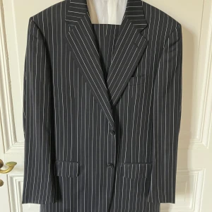 Kritstrecksrandig blå kostym från Corneliani - Superelegant mörkblå kostym från Corneliani i klassiskt kritstrecksrandigt mönster. Kavajen har breda slag, två knappar och fickor med lock. Byxorna matchar med samma randiga stil. Tillverkad i exklusiv extrafin merinoull Super 120’s. Perfekt för dig som vill sticka ut med stil.