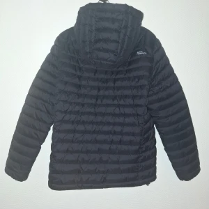 Svart pufferjacka från 157 med huva - Svart pufferjacka från 157 med huva och dragkedja framtill. Jackan har quiltad design med horisontella sömmar och två fickor på sidorna. Perfekt för kyliga dagar och enkel att matcha med det mesta.