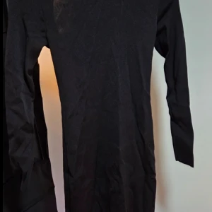 Svart långärmad klänning med snörning - Svart långklänning med långa ärmar och snygg snörningsdetalj i ryggen. Klänningen har en tight passform och är tillverkad i ett stretchigt material som sitter skönt mot kroppen. Perfekt för dig som gillar minimalistisk stil med en twist.