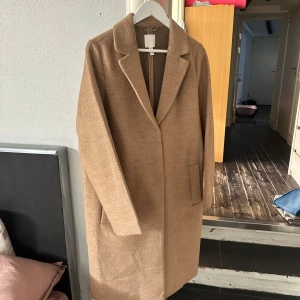 Beige kappa från H&M - Stilren beige kappa från H&M med klassisk krage och raka linjer. Jackan har två fickor framtill och är helfodrad. Perfekt för dig som gillar en clean och tidlös look. Materialet är mjukt och känns lyxigt, passar perfekt till höst och vår.