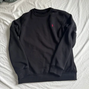 Svart sweatshirt från Ralph Lauren - Svart sweatshirt från Ralph Lauren med klassisk rund hals och broderad rosa logga på bröstet. Tröjan har ribbade muddar vid ärmslut och nederkant, samt långärmad passform. Perfekt för en clean och stilren look.