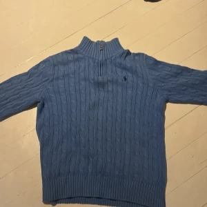 Blå kabelstickad tröja Polo Ralph Lauren - Kabelstickad blå tröja från Polo Ralph Lauren med halv dragkedja i halsen och broderad logga på bröstet. Tröjan har ribbade muddar och hög krage med svarta ränder på insidan. Perfekt för kyliga dagar.