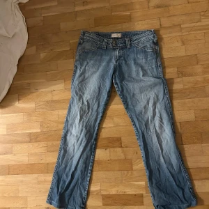 Blå jeans från Miss Sixty - Snygga blå jeans från Miss Sixty med klassisk femficksmodell och dubbla knappar i midjan. Jeansen har raka ben och en låg midja. Materialet är klassisk denim i bomull med slitningar för en cool look. Midjemåttet är 38cm