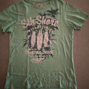 Grön t-shirt från Sth Shore - Snygg grön t-shirt från Sth Shore med tryck av surfbrädor, palmer och texten 'Ipanema Beach'. Skön passform och tillverkad i mjuk bomull. Perfekt för en avslappnad stil med surfvibbar.