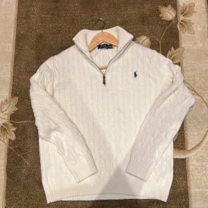 Vit kabelstickad tröja Polo Ralph Lauren - Kabelstickad vit tröja från Polo Ralph Lauren med halv dragkedja och klassisk blå logga på bröstet. Tröjan har hög krage och långa ärmar, perfekt för kyliga dagar. Snygg och tidlös design som passar till det mesta. Helt Oanvänd
