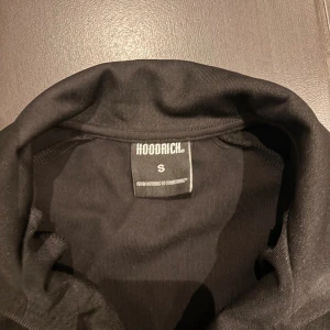 Svart ziptröja från Hoodrich S - Svart ziptröja från Hoodrich med vit logga på bröstet och längs båda ärmarna. Tröjan har hel dragkedja, ståkrage och ribbade muddar. Tillverkad i ett mjukt och lätt material som passar perfekt till streetwear-stilen.
