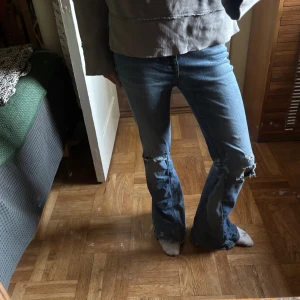 Blå bootcut jeans med slitna knän - Utsvängda jeans med hål vid knäna. De har normal hög midja och är omsydda till bootcut nere vid benen, vilket gör dom väldigt coola. Storlek står inte men sitter som XS