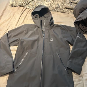 Svart Haglöfs Gore-Tex vindjacka L - Säljer en svart vindjacka från Haglöfs i storlek L. Jackan är tillverkad i slitstark Gore-Tex med huva, hel dragkedja och två snedställda fickor med dragkedja. Perfekt för blåsiga och regniga dagar, stilren och funktionell design . HELT NYTT