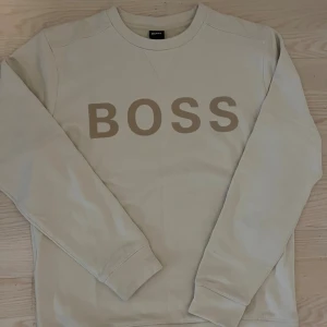 Beige sweatshirt från Hugo Boss - Snygg beige sweatshirt från Hugo Boss med stort BOSS-tryck framtill. Tröjan har rund halsringning och långa ärmar, perfekt för en clean och stilren look. Tillverkad i mjuk bomull för maximal komfort.