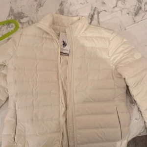 Vit pufferjacka från U.S. Polo Assn. - Snygg vit pufferjacka från U.S. Polo Assn. med diskret logga på ärmen och klassisk quiltad design. Jackan har dragkedja framtill och två fickor med dragkedja. Perfekt för kyliga dagar och ger en clean look. Köpt utomlands!!! Använd en gång!! Etiketten finns inte med!! 
