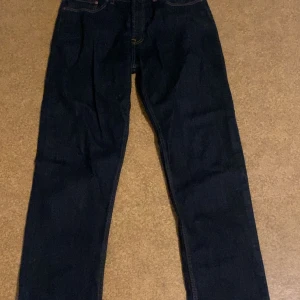 Mörkblå jeans från Dressmann 33/32 - Säljer ett par klassiska mörkblå jeans från Dressmann i storlek 33/32. Jeansen har raka ben, fem fickor och snygga kontrastsömmar. Perfekta för dig som gillar en enkel och stilren look.