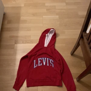 Röd hoodie från Levi's med tryck - Röd hoodie från Levi's (äkta)med stort vitt och blått tryck på bröstet. Klassisk modell med huva och långa ärmar, insida av huvan i vitt. Perfekt för dig som gillar streetwear och vill ha en snygg och bekväm tröja.