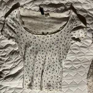 Vit blommig topp med spets och rosett - Supersöt vit topp med små blå blommor, ribbad struktur och korta ärmar. Urringningen är dekorerad med spetskant och en liten rosett framtill. Perfekt för en romantisk och söt look, passar till både jeans och kjol.