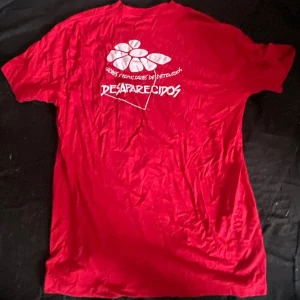Röd t-shirt från AE Promo XL - Röd t-shirt från AE Promo i storlek XL med tryck på ryggen som säger 'Desaparecidos' och har en vit blomma. Klassisk passform och korta ärmar, tillverkad i mjuk bomull. Perfekt för dig som gillar statement-plagg och vill sticka ut.