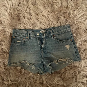 Blå jeansshorts från Perfect Jeans - Snygga blå jeansshorts från gina med råa kanter och slitna detaljer framtill. Klassisk femficksmodell med knapp och dragkedja. Perfekta för sommaren och ger en avslappnad vibe till din outfit.