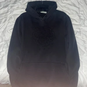 Svart Kenzo hoodie med broderad tiger - Svart hoodie från Kenzo med ikonisk broderad tiger och Kenzo Paris-logga på bröstet. Klassisk huva, känguruficka och mjukt material som känns skönt mot huden. Perfekt för dig som vill ha en stilren och trendig look.