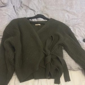 Olivgrön omlottkofta från Moocci - Säljer en olivgrön stickad omlottkofta från Moocci med knytband i midjan och djup v-ringning. Koftan har långa ärmar och grovstickad struktur som ger en mysig och trendig look. Perfekt att slänga på sig under kyliga dagar.