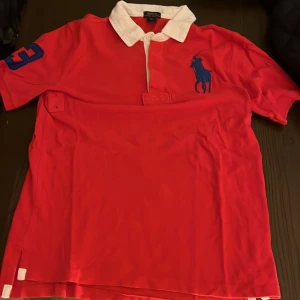 Röd pikétröja från Polo Ralph Lauren - Röd pikétröja från Polo Ralph Lauren med vit krage och blå logga på bröstet. Tröjan har korta ärmar, blå siffra 3 på ena ärmen och klassisk knappslå framtill. Perfekt för en sportig och stilren look.