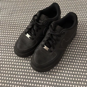 Svarta Nike Air Force 1 sneakers - Klassiska helsvarta Nike Air Force 1 sneakers i skinn med perforerad tå, rund tå och platt sula. Snygg och tidlös modell med snörning och metallbricka med 'AF1' på snörena. Perfekta för dig som gillar streetstyle och vill ha en ikonisk sneaker i garderoben.