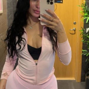 Ljusrosa zip hoodie i sportig stil - Säljer en ljusrosa hoodie med dragkedja och lång ärm. Tröjan har en tight passform och är tillverkad i ett stretchigt material, perfekt för träning. Tumhål vid ärmslut ger extra sportig känsla. Enkel och snygg design som passar till gymmet.