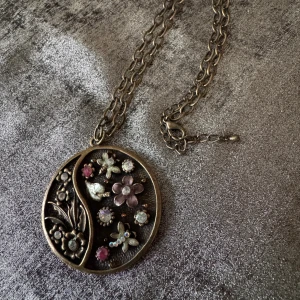 Vintage halsband med blommor och stenar - Unikt halsband med rund medaljong i antikguldfärg. Framsidan har blommor och små gnistrande stenar i lila, rosa och silver, samt detaljerade blad och fjärilar. Kedjan är grovlänkad och medaljongen har en tydlig boho-vibe. Perfekt för dig som gillar detaljer och vintagekänsla.medaljongen är ca5 cm i diameter.