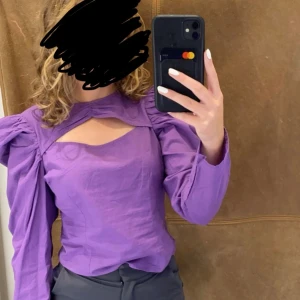 Lila blus med puffärmar och cut out - Säljer en lila blus med puffärmar och cool cut out-detalj både fram och bak. Blusen har lång ärm, figurnära passform och knappar i nacken. Perfekt för dig som vill sticka ut med en trendig och unik topp på en utekväll! Aldrig använd!