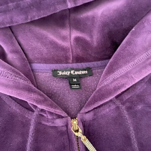 Lila vintage hoodie Juicy Couture  - Juicy Couture hoodie i lila velour med dragkedja och glittrande J-hänge. Klassisk huva, fickor framtill. Perfekt för dig som vill ha den ikoniska 2000-talslooken med extra bling.