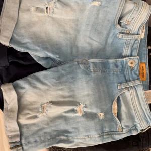 Ljusblå jeansshorts Jack & Jones - Snygga ljusblå jeansshorts från Jack & Jones med slitna detaljer och upprullade benslut. Klassisk femficksmodell med knapp och dragkedja. Perfekta för en avslappnad stil under varma dagar.
