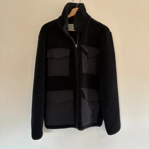 Svart teddyjacka från A Day's March - Svart teddyjacka från A Day's March med fyra stora fickor framtill och dragkedja. Jackan har hög krage och är tillverkad i mjuk fleece med detaljer i syntetmaterial. Perfekt för dig som gillar en clean och stilren look. Finns liten spricka på insidan av kragen vilken syns på sista bilden.