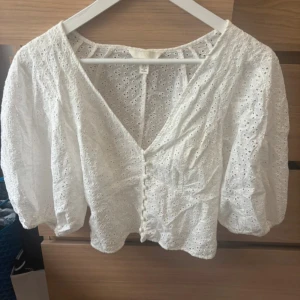Vit broderad blus från H&M - Superfin vit blus från H&M i broderad bomull med v-ringning, knappar framtill och puffiga trekvartsärmar. Croppad modell med romantiska broderidetaljer och luftig känsla. Perfekt till jeans eller kjol för en söt look.