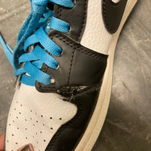 Nike Jordan 1 svart/vit med blå detaljer - Säljer ett par Nike Jordan 1 sneakers i svart och vitt skinn med blå snörning och blå vadderad insida. Klassisk design med perforerad tå och svart swoosh på sidan. Perfekta för dig som gillar streetwear och vill sticka ut med färgglada detaljer. Obs- mycket väl använda. Defekter förekommer inuti såväl som utanpå. Se bilder. Funkar dock att bära skorna utan problem 