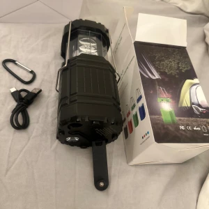 PS-201 Solar Hand Crank Camping Lantern solcellslampa - Multifunktionell campinglampa med solcellsladdning, handvev och USB-port. Robust design, perfekt för utomhusbruk. Levereras med karbinhake och laddkabel. Produkten ser oanvänd ut och levereras i originalförpackning. Inga synliga defekter eller slitage.