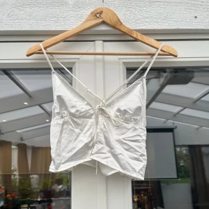 Vit satinlinnetopp med knytning - Snygg vit linnetopp i satin med smala axelband och knytning framtill. Toppen har en lätt glansig finish och spetsig nederkant, vilket ger en trendig och feminin look. Perfekt för att styla med jeans eller kjol för en cool vibe.