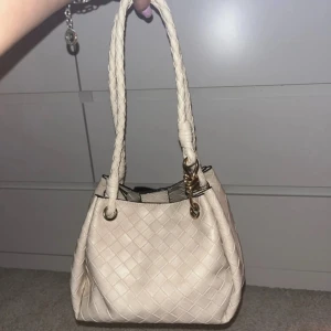 Beige flätad handväska med guldiga detaljer - Snygg beige handväska i flätat skinnimitation med två rejäla handtag och guldiga metalldetaljer. Väskan har ett rymligt fack och en matchande innerpouch med dragkedja. Perfekt för dig som gillar stilrena accessoarer med lite extra edge.