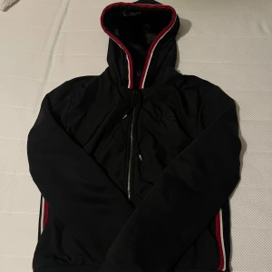 Dolce & Gabbana Cardigan - Svart vindjacka från D&G med huva och dragkedja framtill. Jackan har röda och vita ränder längs huva och sidor, samt diskret D&G-logga på bröstet. Perfekt för dig som vill ha en stilren och sportig look. Storlek 46/S, nypris 7000kr säljs för 2000kr