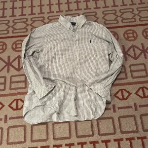 Rutig skjorta från Ralph Lauren - Klassisk vit skjorta från Ralph Lauren med mörkblå rutor och broderad logga på bröstet. Skjortan har lång ärm, knappar framtill och klassisk krage. Classic fit-modell i mjuk bomull, perfekt för en clean och stilren look.