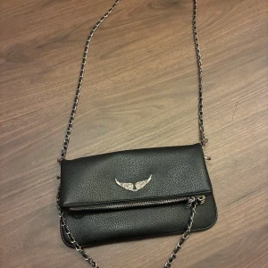 Svart axelväska med kedjerem - Snygg svart axelväska från Zadig & Voltaire i skinn med silverfärgad kedjerem och dragkedjedetaljer. Framsidan har en ikonisk vinglogga i metall. Perfekt storlek för det viktigaste. Väskan har lite sminkfläckar i botten annars i fint skick!