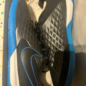 Nike svarta och blå futsalskor (Nike tiempo futsal) - Säljer ett par grymma Nike fotbollsskor med svart ovandel i syntetmaterial och coolt diamantmönster. Skorna har snörning och en blå yttersula med bra grepp, samt Nike-logga med blå kant. Perfekt för dig som vill sticka ut på planen.