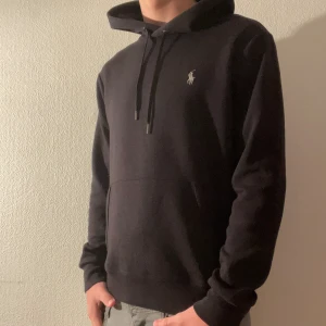 Svart hoodie från Polo Ralph Lauren - Svart hoodie från Polo Ralph Lauren med klassisk broderad logga på bröstet. Tröjan har huva med snörning, känguruficka och långa ärmar. Perfekt för en chill och stilren look. Modellen är 176 och väger cirka  65kg