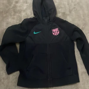 Svart Nike FC Barcelona hoodie 137-148cm - Svart hoodie från Nike med FC Barcelona-logga i rosa och turkos Nike-swoosh på bröstet. Tröjan har huva, dragkedja framtill och två fickor. Perfekt för dig som älskar fotboll och vill ha en sportig look.