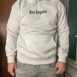 Vit hoodie från shein  - Vit hoodie med svart Los Angeles-tryck på bröstet och stort vertikalt tryck på ryggen. Har huva och känguruficka framtill samt tryckt text på ena ärmen. Perfekt för dig som gillar streetstyle och vill sticka ut.