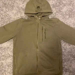 Olivgrön Sail Racing hoodiejacka - Olivgrön hoodiejacka från Sail Racing med dragkedja framtill, bröstficka med dragkedja och broderad logga. Jackan har huva med snörning och mjukt fleecefoder på insidan. Perfekt för kyliga dagar och snygg streetstil.