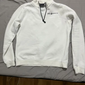 Vit Sail Racing half zip tröja - Vit långärmad tröja från Sail Racing med svart half zip-dragkedja och logga på bröstet. Tröjan har hög krage och är tillverkad i mjukt material, perfekt för kyliga dagar. Enkel och stilren design som passar till det mesta. 