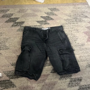 Svarta cargo bermudashorts Big Star - Svarta bermudashorts från Big Star med cargofickor på sidorna och klassisk jeanslook. Modellen heter Morris Bermudas och har flera praktiska fickor samt knapp- och dragkedjestängning. Perfekta för en avslappnad streetstil.