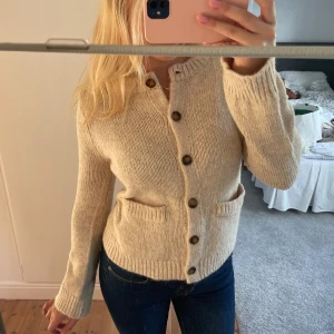 Beige stickad kofta med knappar - Supermysig beige stickad kofta med bruna knappar och två fickor framtill. Koftan har långärmad passform och ribbade muddar vid ärmslut och nederkant. Perfekt för lager-på-lager och chill höst- och vinterstil. Helt oanvänd!