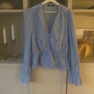 Ljusblå blus med broderade detaljer - Supersöt ljusblå blus från Bik Bok med broderade hålmönster och volangkant. Blusen har v-ringning, knappar framtill och långa ärmar med rynk och volang vid ärmslut. Nästan aldrig använd!
