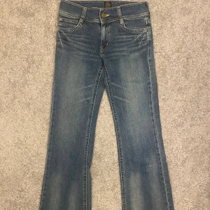 Blå bootcut jeans med fickdetaljer - Snygga blå jeans med bootcut passform och coola bakfickor med lock och guldiga knappar. Jeansen har klassisk femficksdesign, kontrastsömmar och lätt slitning framtill. Perfekta för dig som gillar en avslappnad men trendig look. Midja rakt över: 35cm Lår: 24cm Innerben: 78cm 